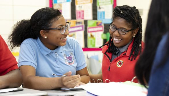 a student smiles and looks at an AmeriCorps member as they sit together at a table doing work