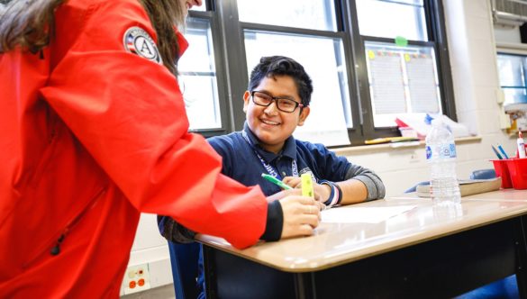 an elementary student is working at desk while AmeriCorps member is standing by to help