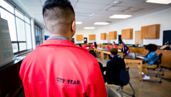 the back of a city year americorp member with the label City Year on his jacket as he looks over a teacher and students in a classroom