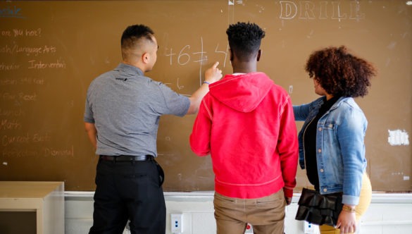 A city year americorps member at a chalkboard showing a math problem to two high school students