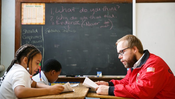 City year americorps member reading along with two students as they sit at desks in a classroom