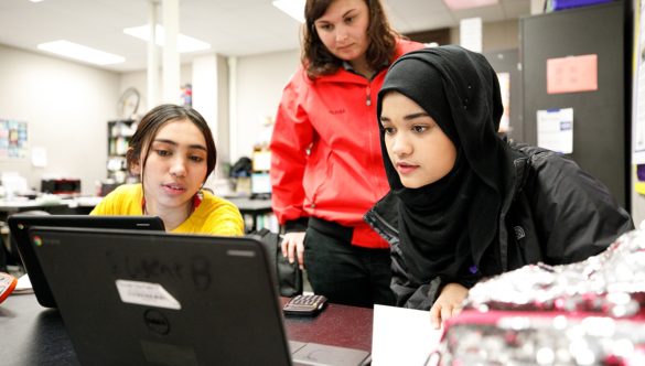 AmeriCorps member stands behind two students as they work at laptops in the classroom