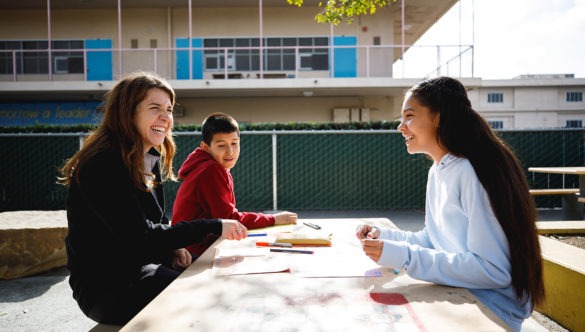 city year americorps member sitting with two students doing work as they sit at a large table outside in the school courtyard