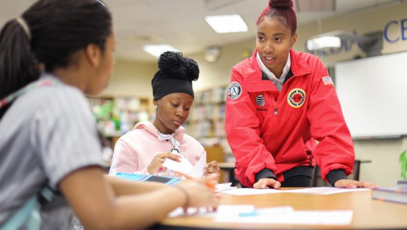 city year americorps member stands and helps two students who are sitting at a table working on schoolwork