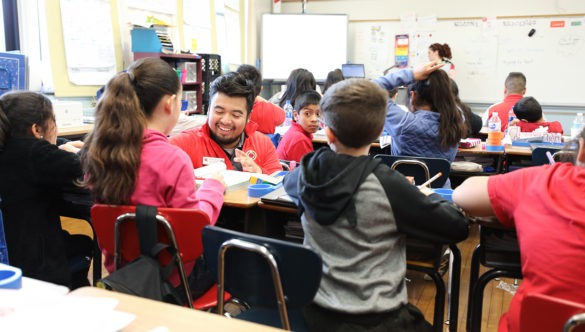 city year americorps member kneeling at a students desk in a full classroom while a teacher teaches