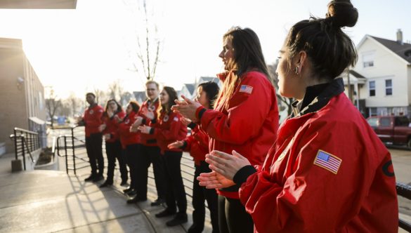 A team of 12 City Year AmeriCorps members stand in a straight line, clapping to welcome their students to school