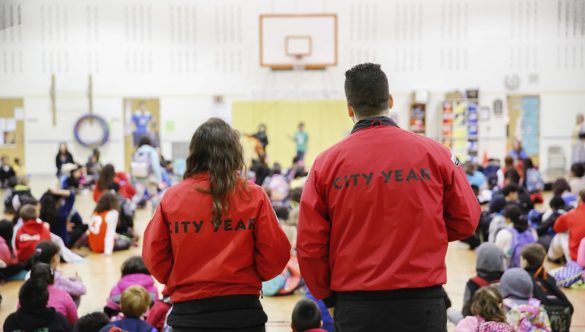two AmeriCorps members are standing behind a large group of students during an assembly in the gym