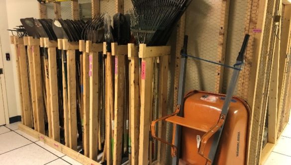 Various landscaping tools in a wooden storage rack. An orange wheelbarrow leans against the wall beside it.